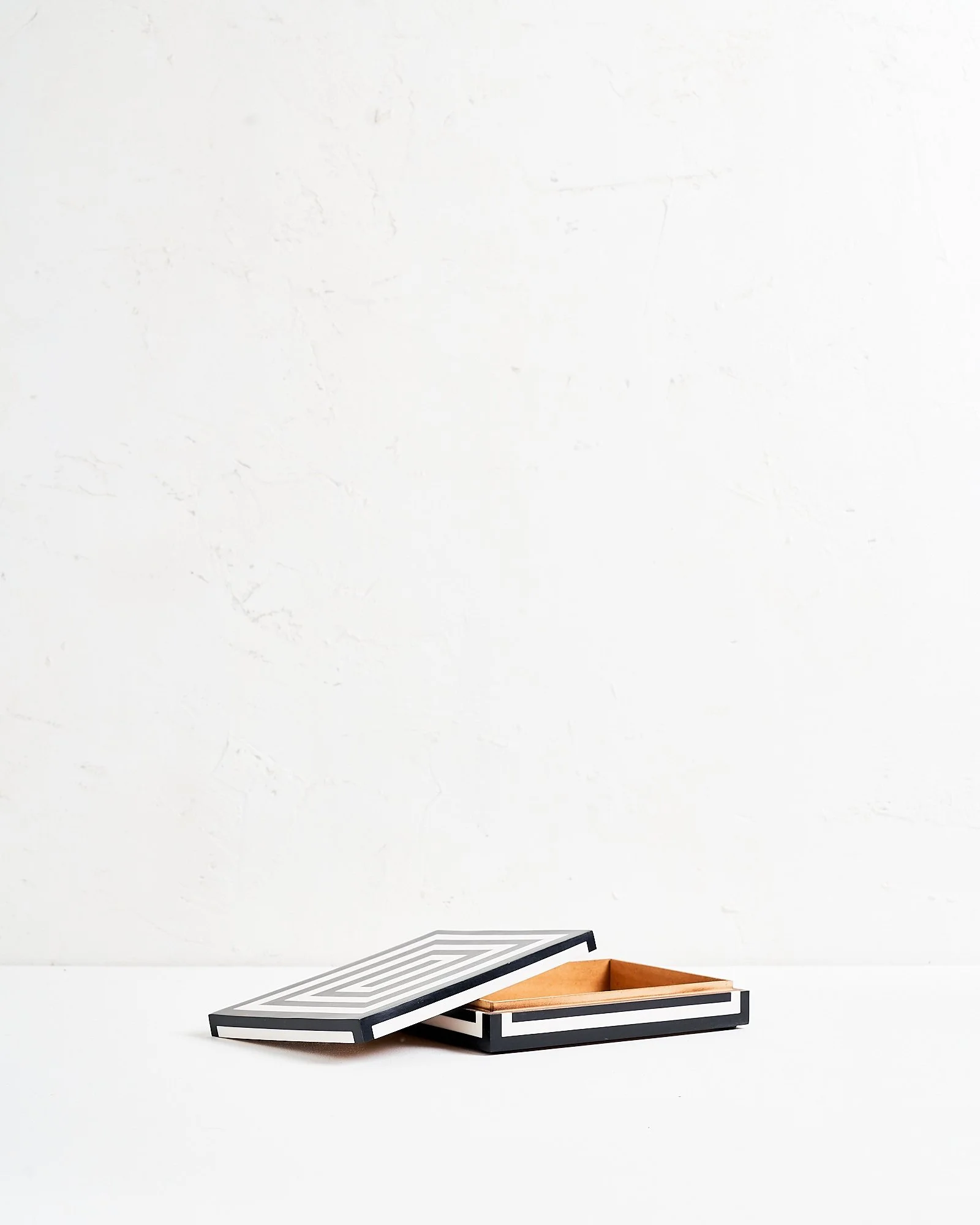 A shallow black and white geometric patterned jewelry box with lid leaning on side on a white surface against a plain white wall.
