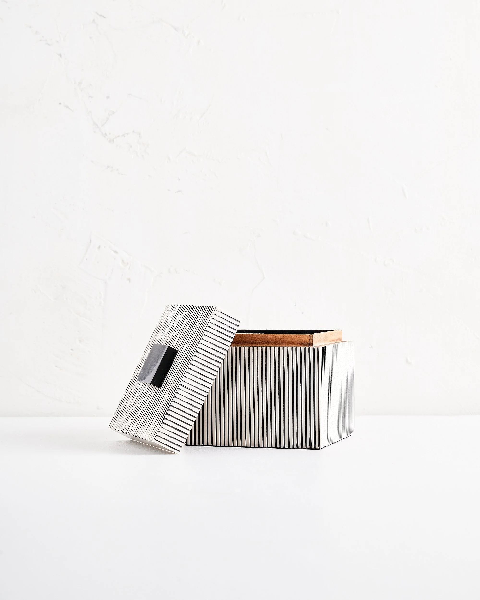 A medium sized modern black and white striped box with a slightly open lid and a black handle on a white surface against a white textured wall.