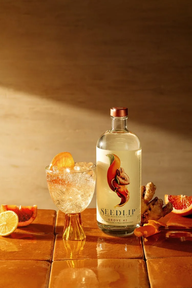 A glass with ice and a grapefruit slice, a bottle labeled 'Seedlip' with a citrus and herbal design, along with sliced lemon, grapefruit, ginger root, and orange peel on a wooden surface.
