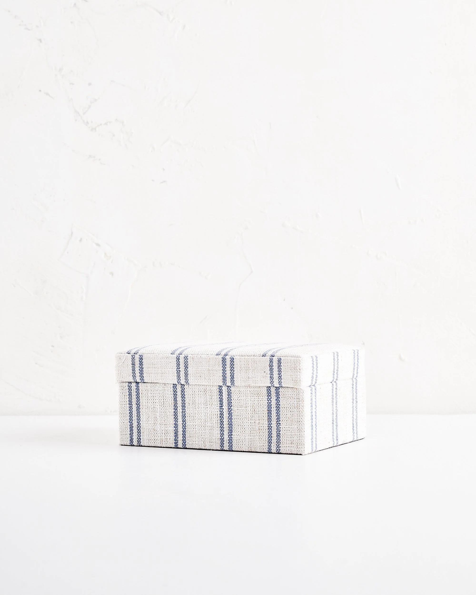 A rectangular fabric-covered box with a blue stripe pattern, placed on a white surface against a white wall.