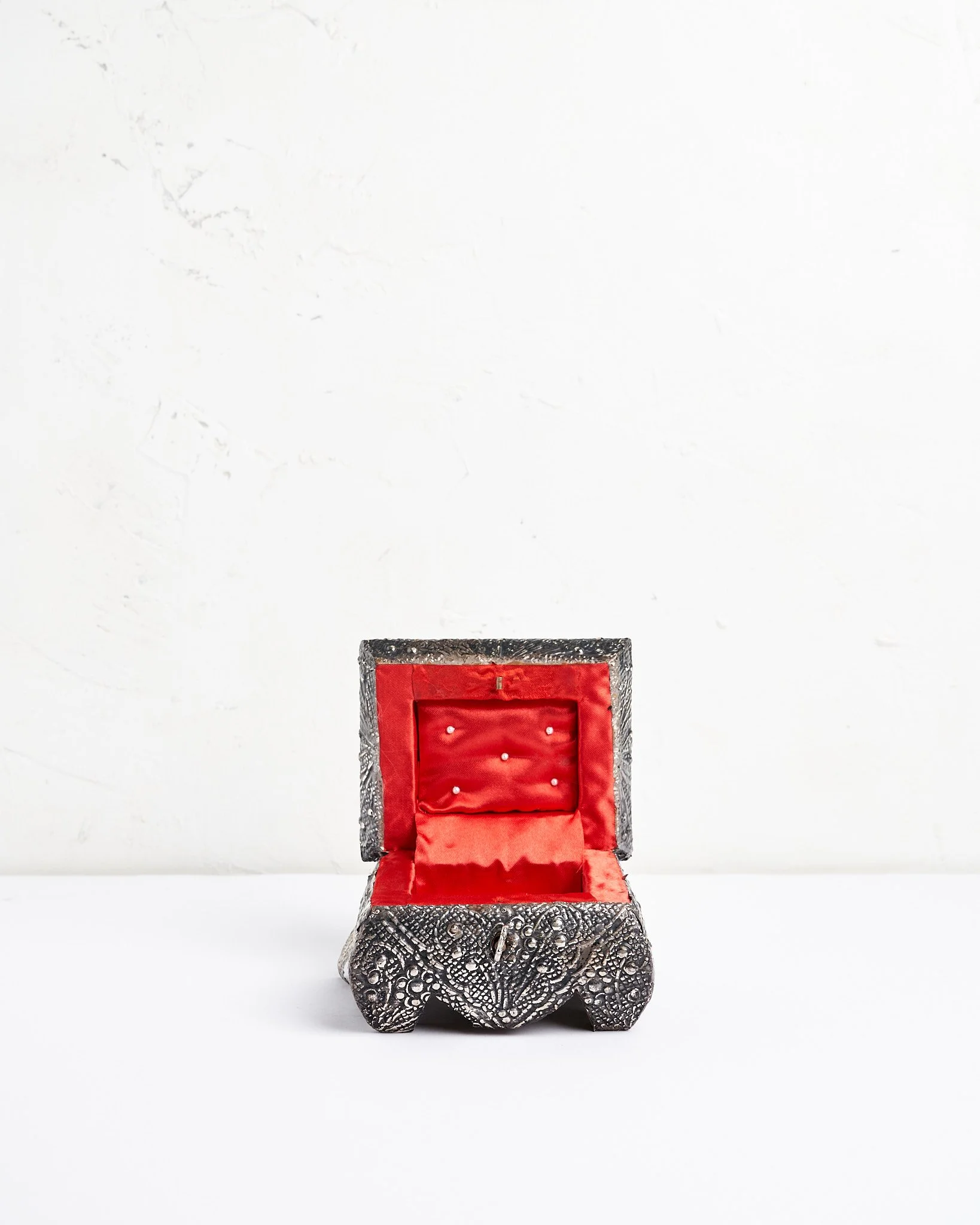 An ornate jewelry box with a textured metallic exterior and a red satin interior, open and placed on a white surface against a plain white background.