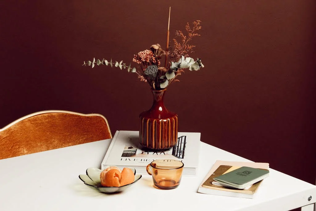 Table with a vase of dried flowers, a glass mug, a bowl with fruit, a closed book, and a magazine, against a dark wall.