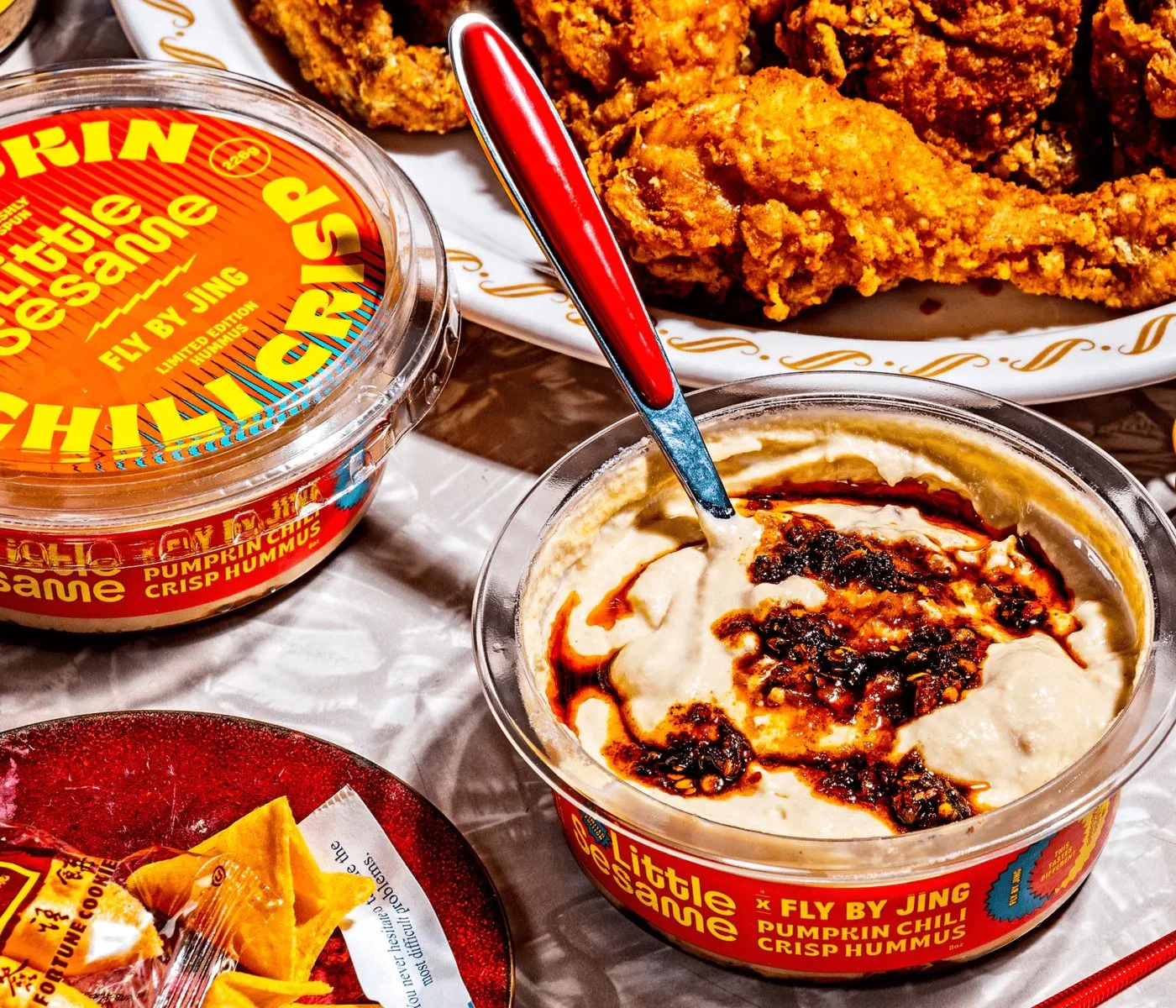 Pumpkin chili crisp hummus by Fly By Jing in a container with a spoon, beside a plate of fried chicken and a small dish with fortune cookies.