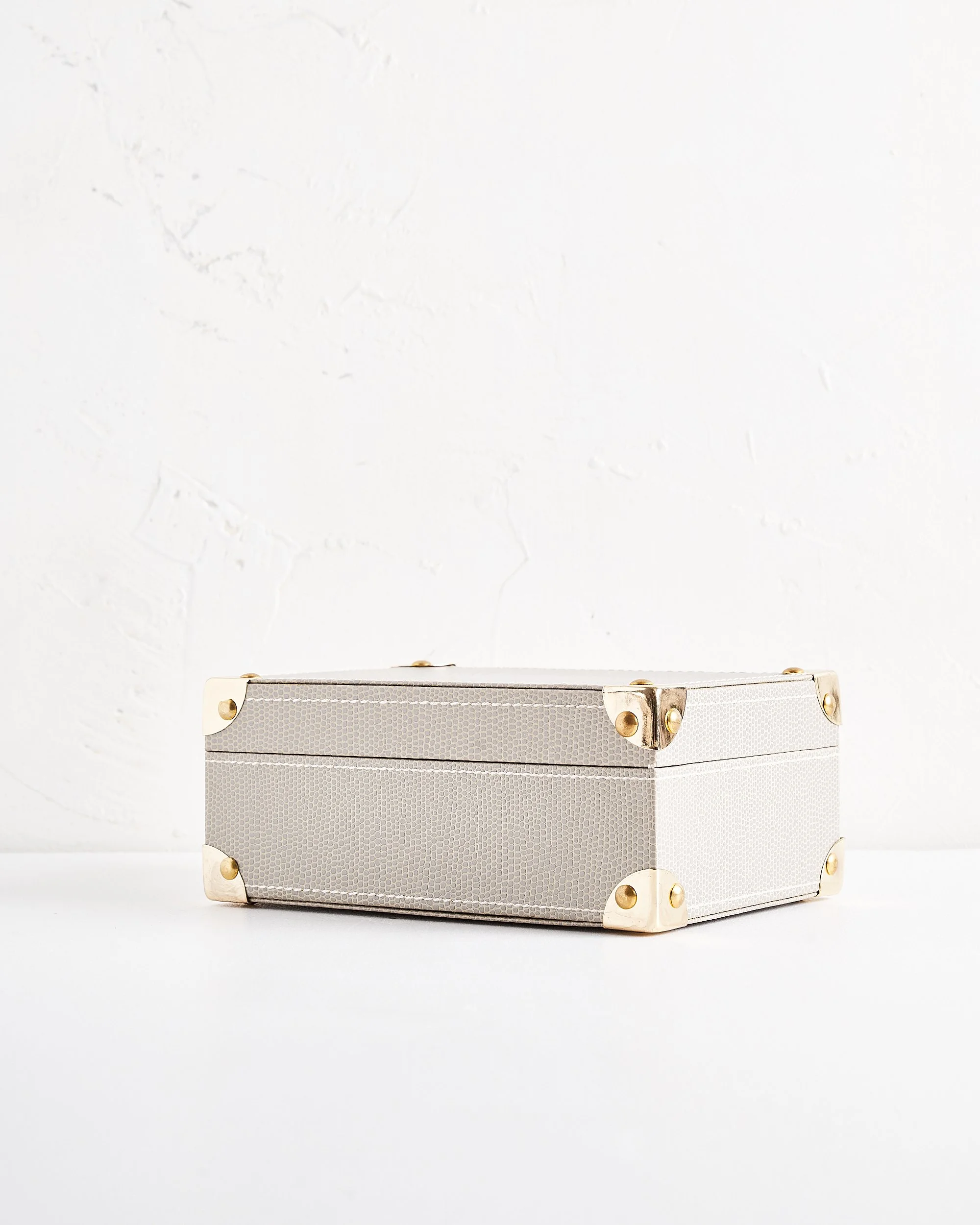 A light gray leather rectangular jewelry box with gold corner accents, placed on a white surface against a textured white wall.