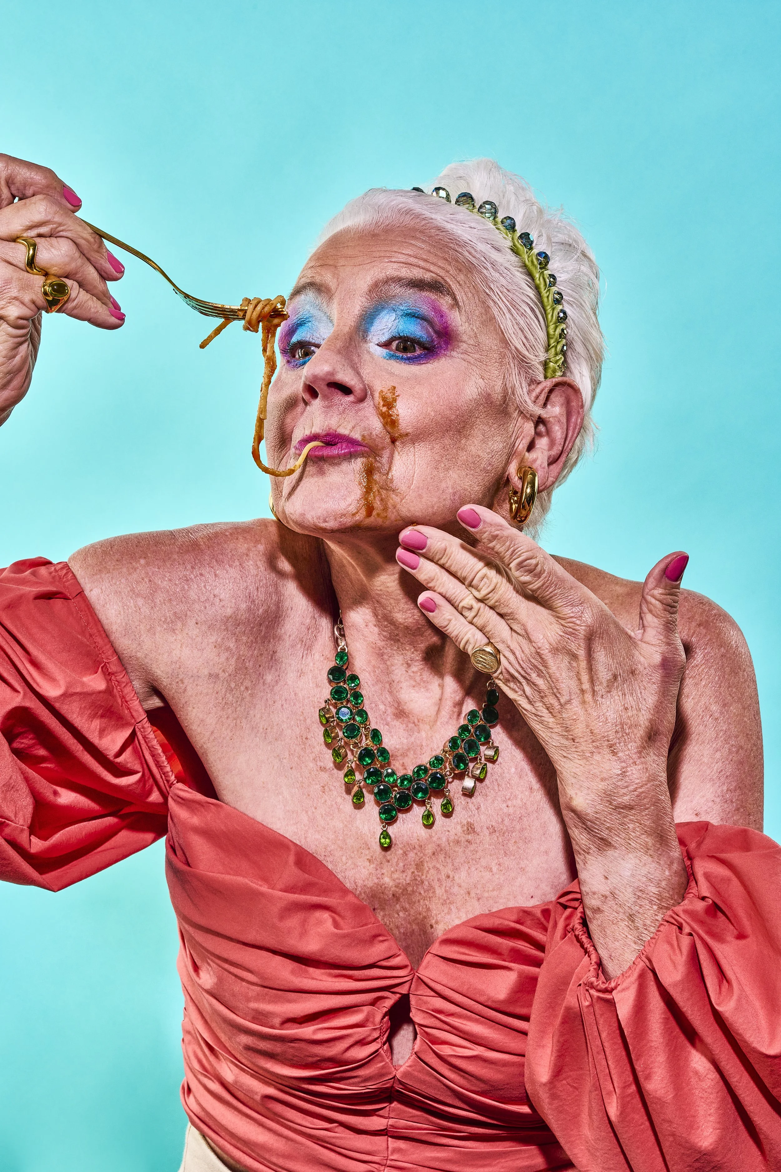 An elderly woman with white hair, wearing colorful makeup, a green jeweled headband, gold earrings, and a green necklace, is eating spaghetti with sauce while posing against a bright blue background.