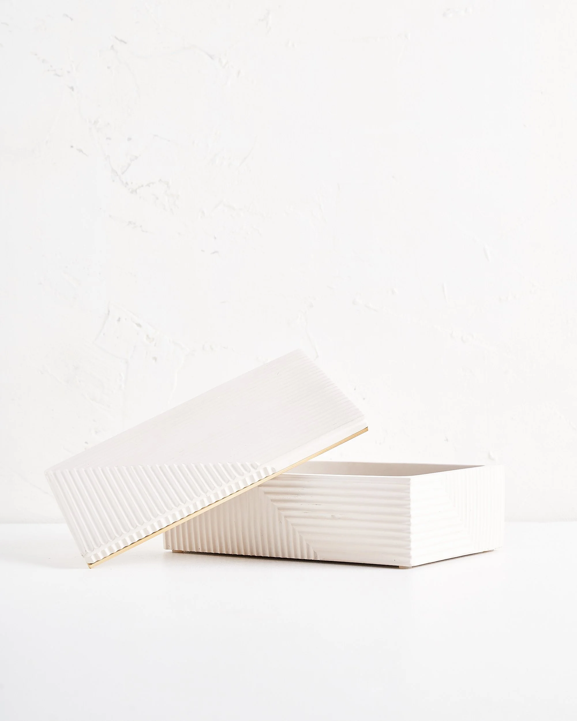 A white modern art deco style white, textured rectangular box with a separate lid, partially open, placed on a white surface against a white wall background.