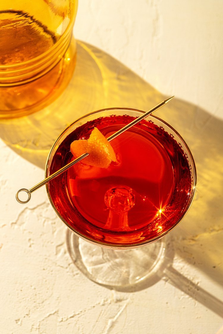 A red cocktail in a stemmed glass garnished with an orange twist on a cocktail pick, with a bright reflection on the surface, placed on a textured white surface, with a yellowish drink and container in the background.