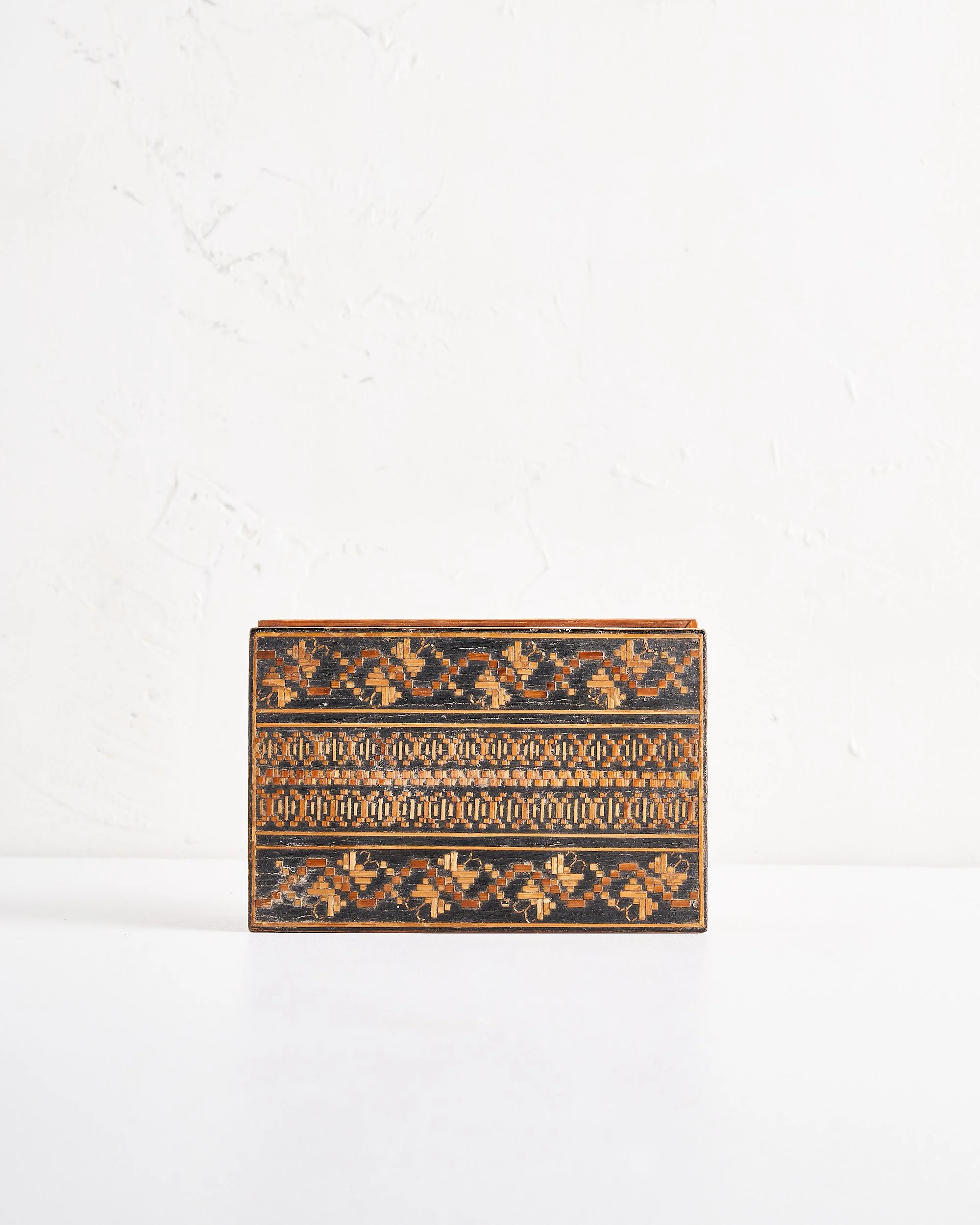 The top of a decorative wooden box with intricate geometric patterns in black, orange, and brown, placed on a white surface against a white wall.