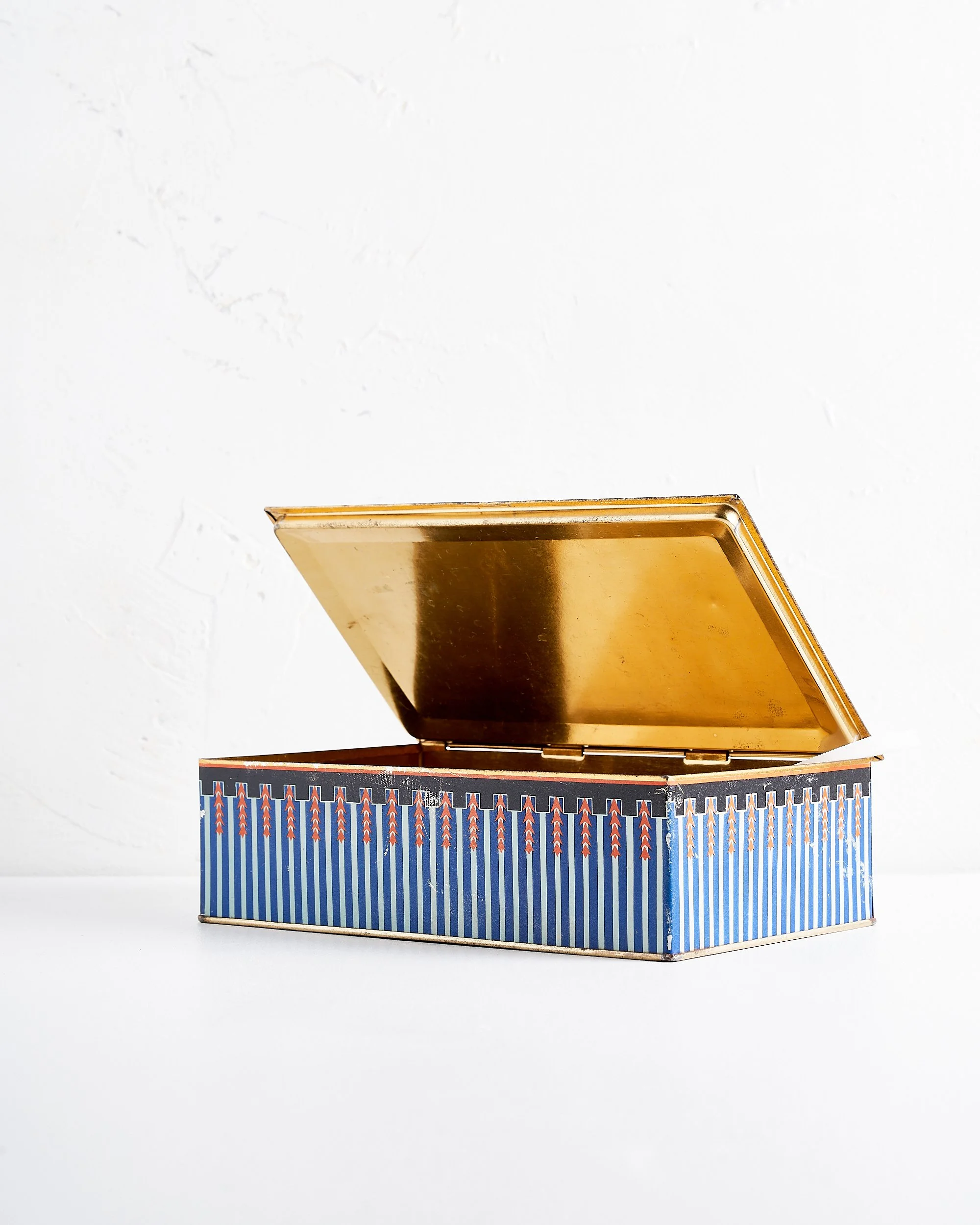 An open vintage rectangular tin box with a decorative patterned exterior lined with gold on the inside, placed on a white surface against a plain white wall.