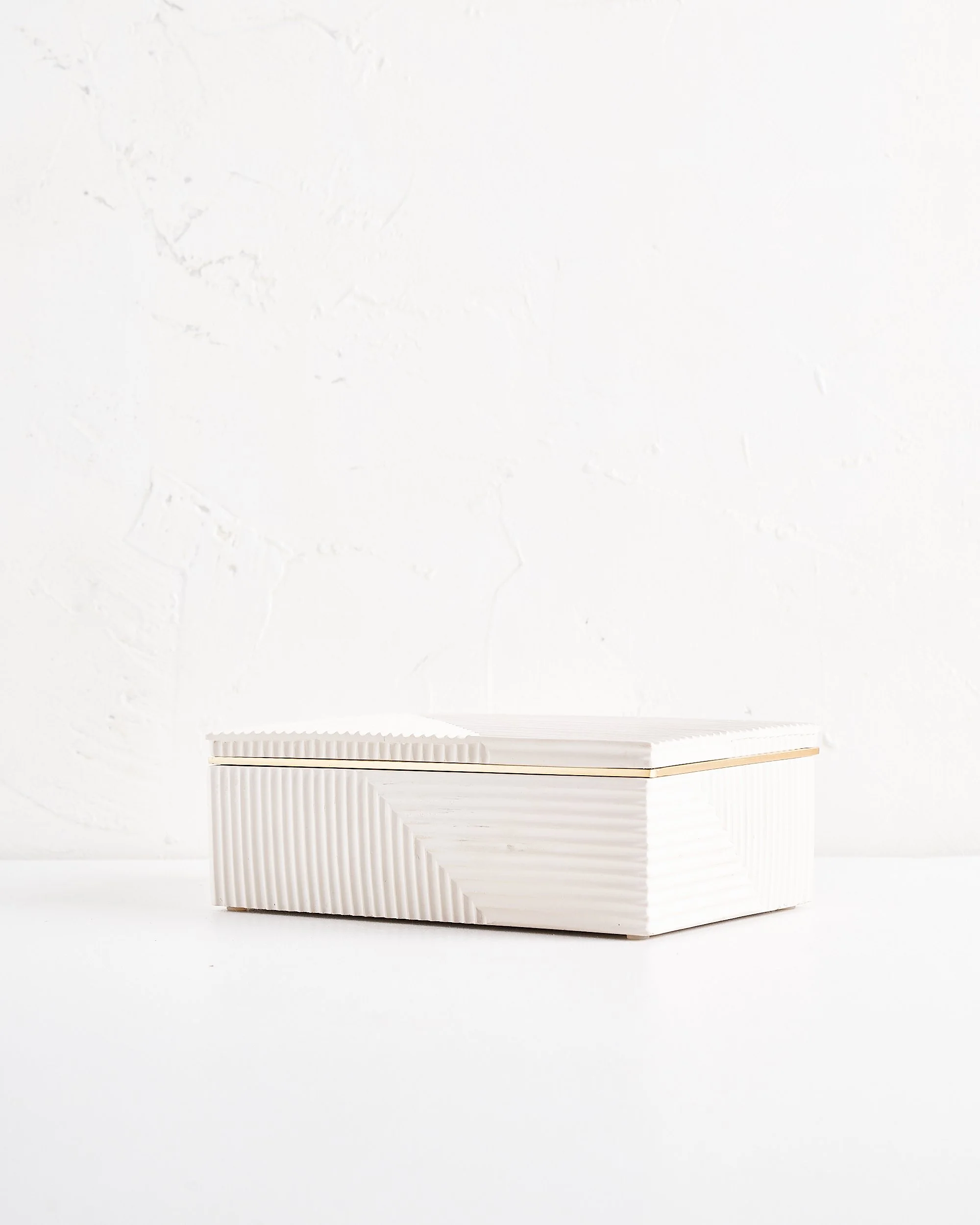 A modern art deco style white, textured rectangular box with a lid, placed on a white surface against a white wall.