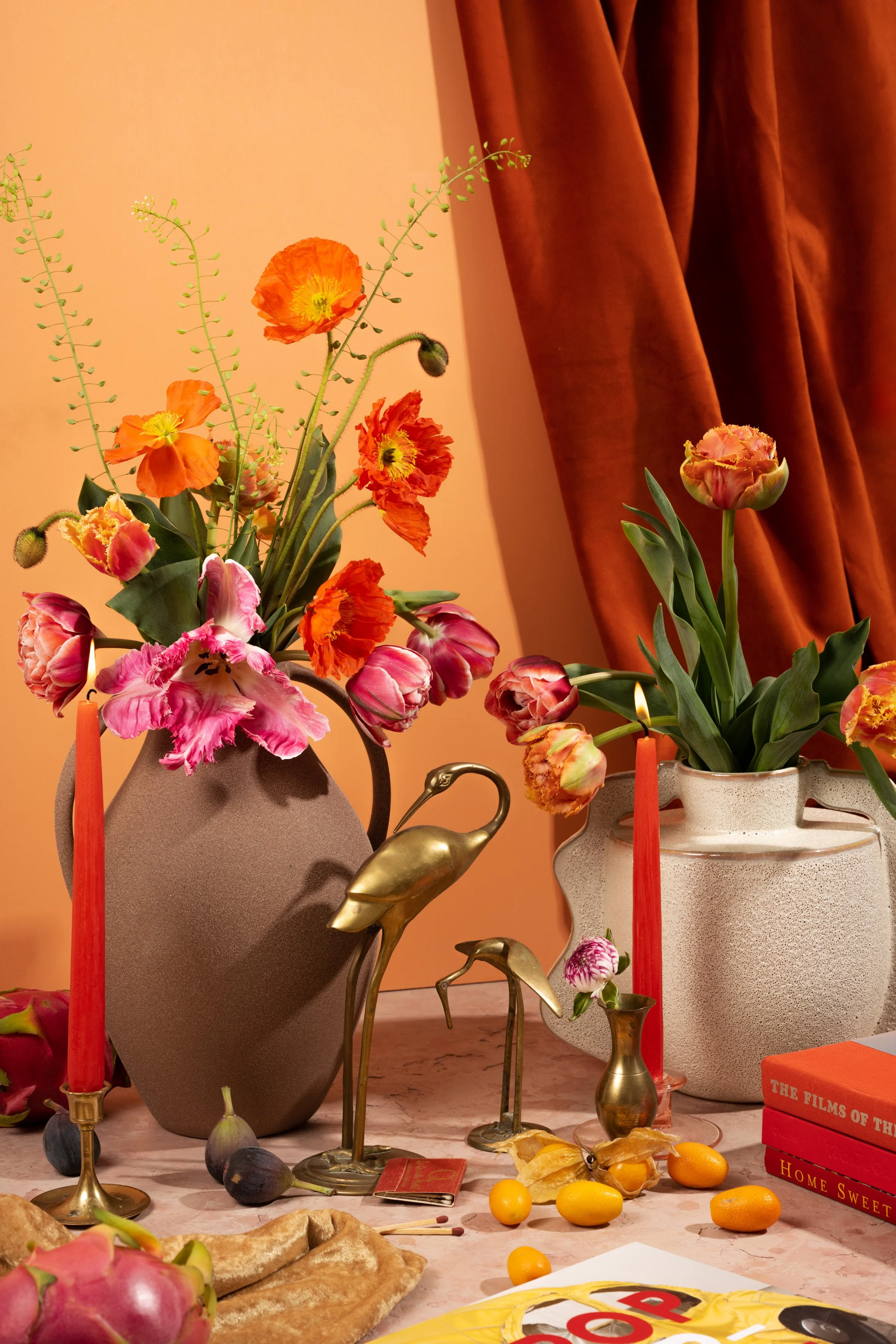 Decorative table arrangement with vases of colorful flowers, including tulips and poppies, two red candles in brass holders, and brass crane figurines. There are also small fruits, a wrapped gift, and books on the table, with a background of orange curtains.