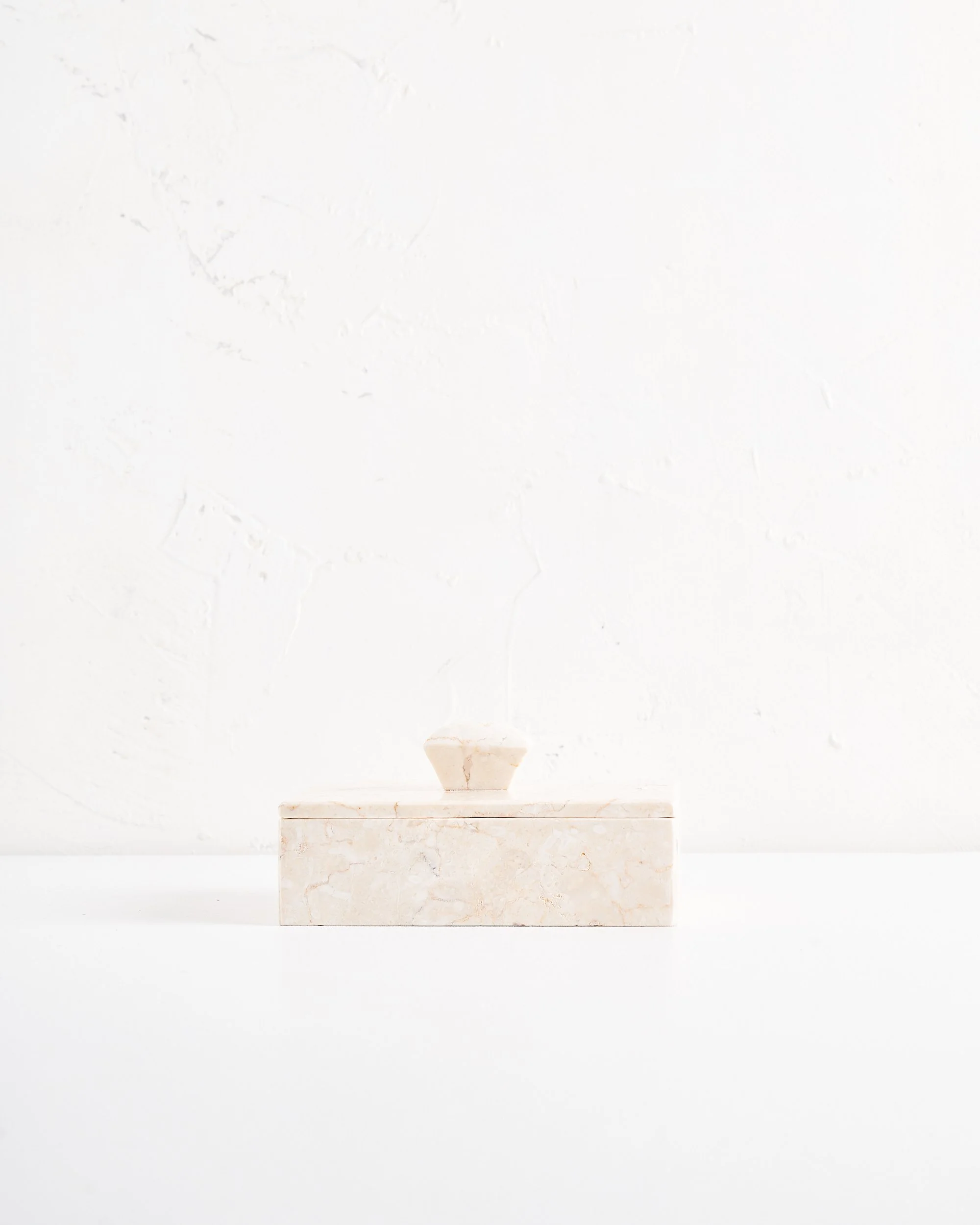 Minimalist and modern light marble jewelry box with handle on lid, sitting on a rectangular marble base against a white textured wall.