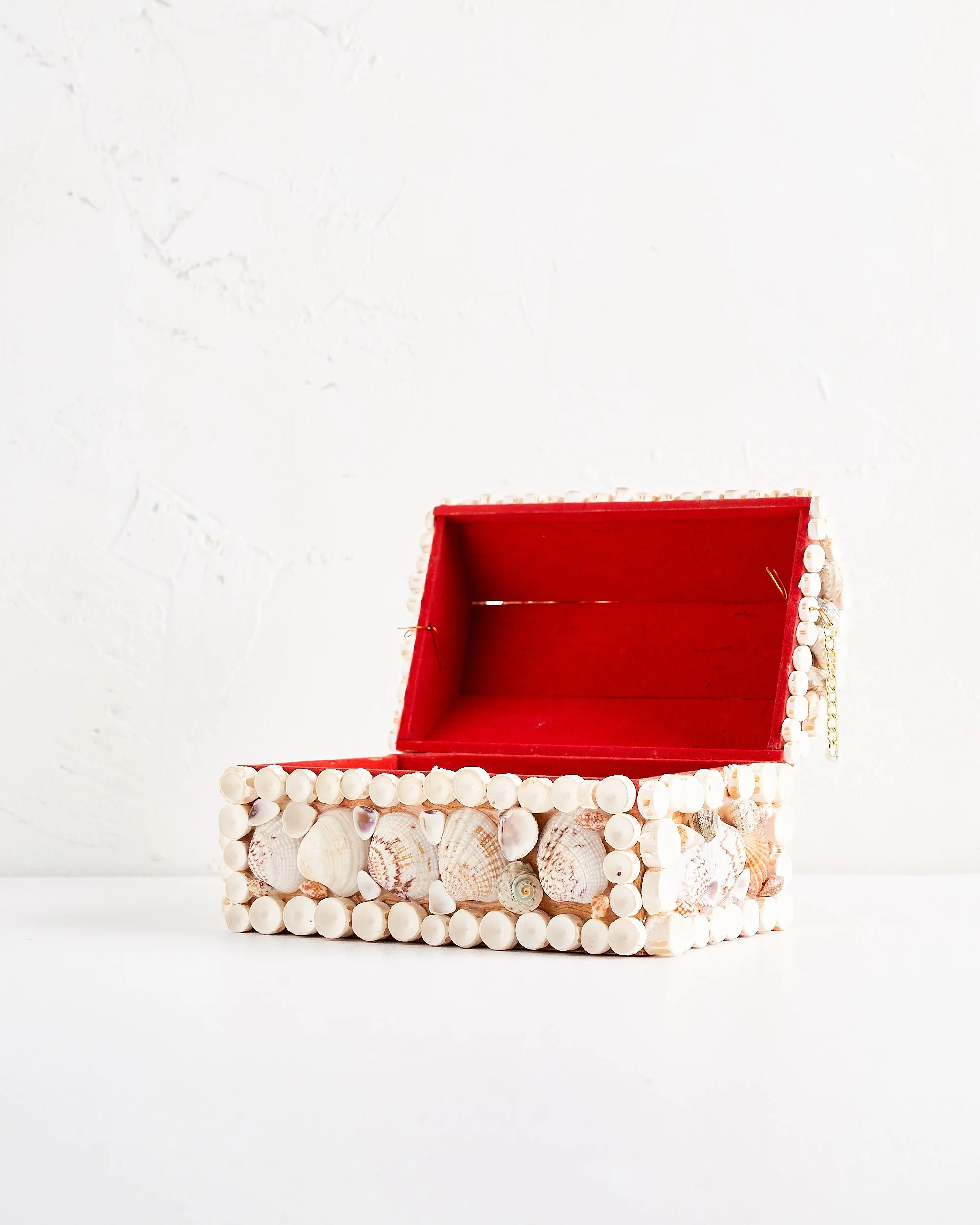 Decorative jewelry box covered with seashells, open to reveal a red interior, placed on a white surface against a textured white wall.
