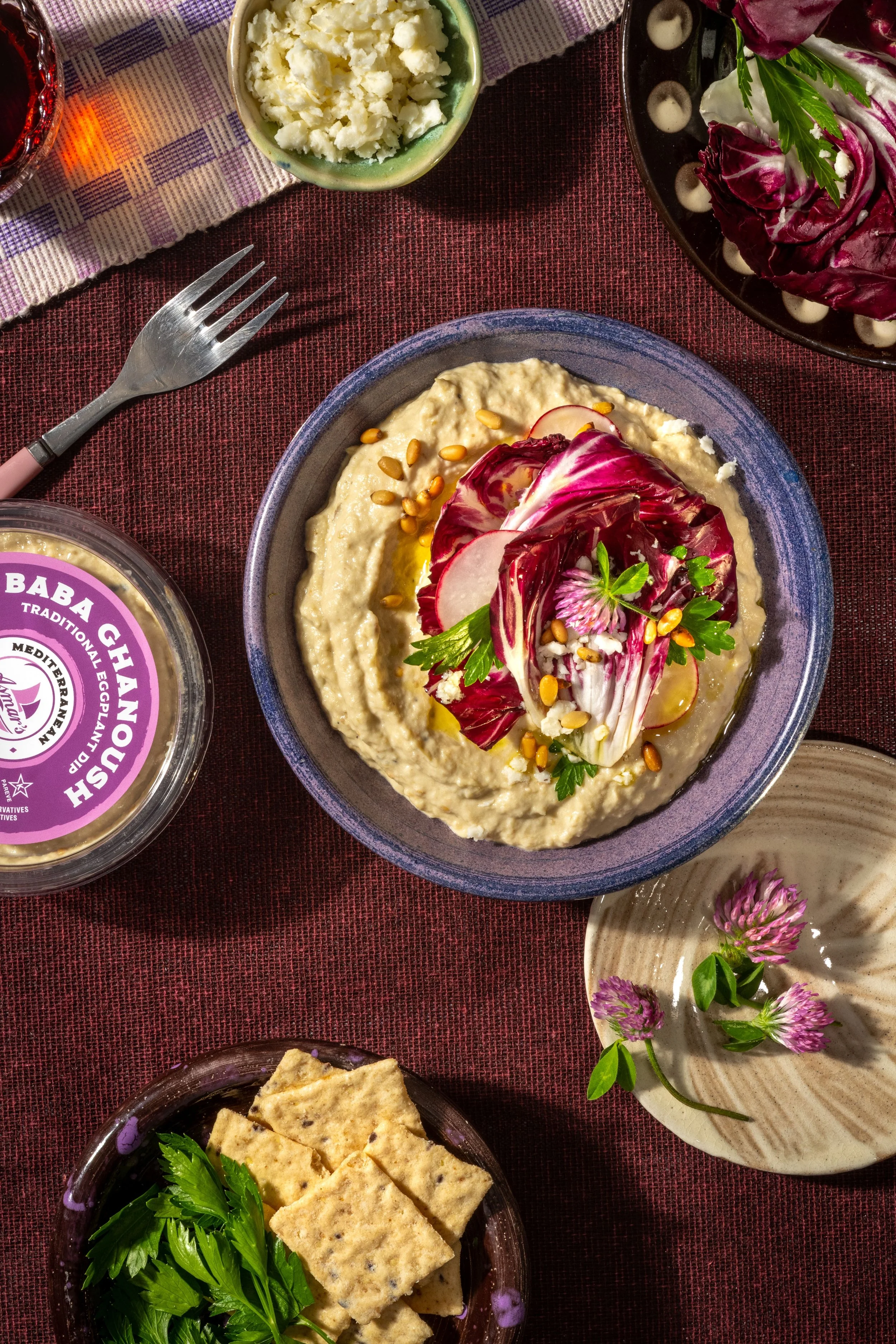 A bowl of hummus topped with radicchio, radish slices, herbs, and pine nuts on a maroon table. Surrounding it are crackers with parsley, a small bowl of crumbled cheese, a plate with edible purple flowers, and a jar of Baba Ghanoush. A fork rests on the table, and a purple, plaid napkin is partially visible.