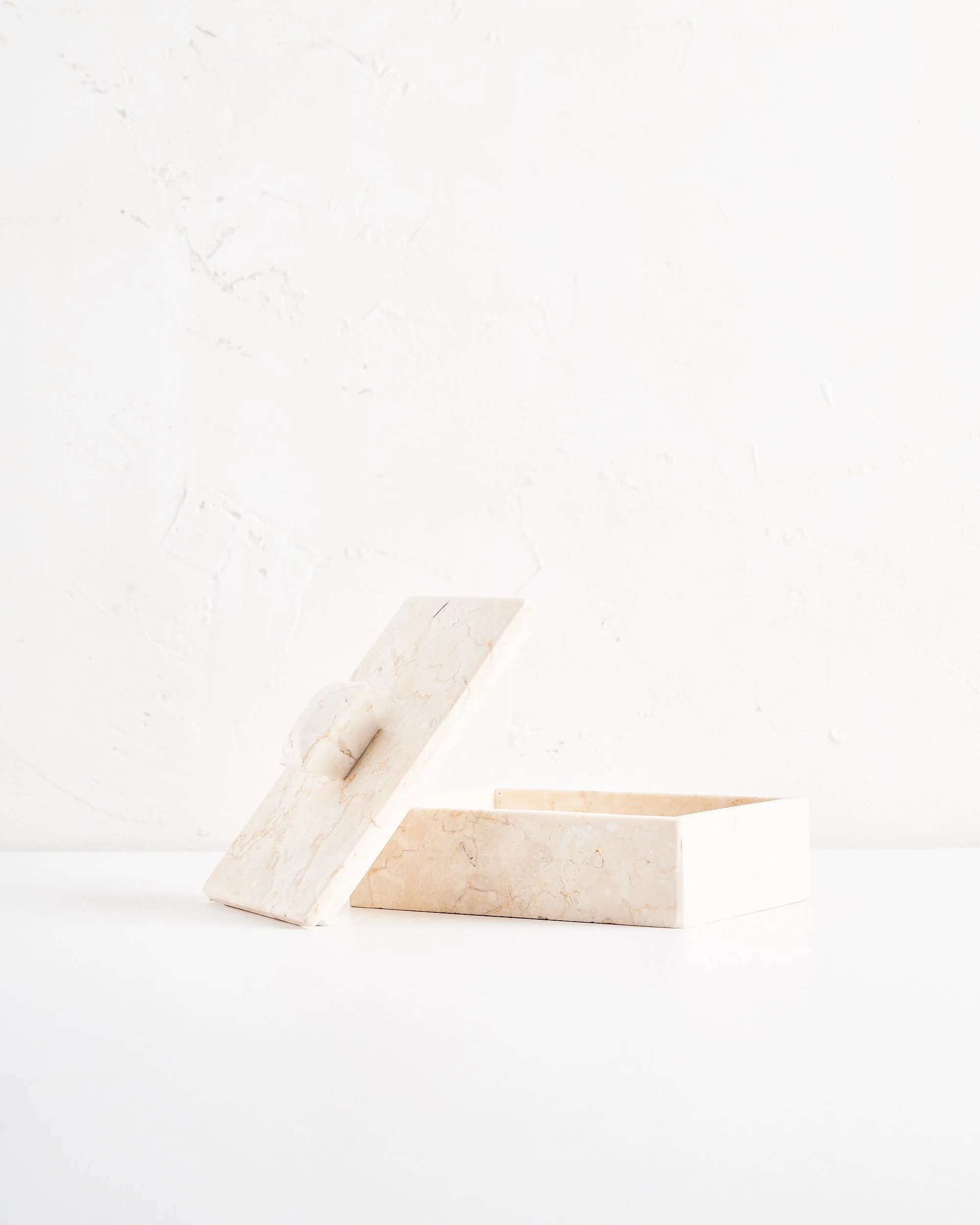 Open modern minimalist light marble jewelry box with lid placed beside it on a white surface against a white textured wall.