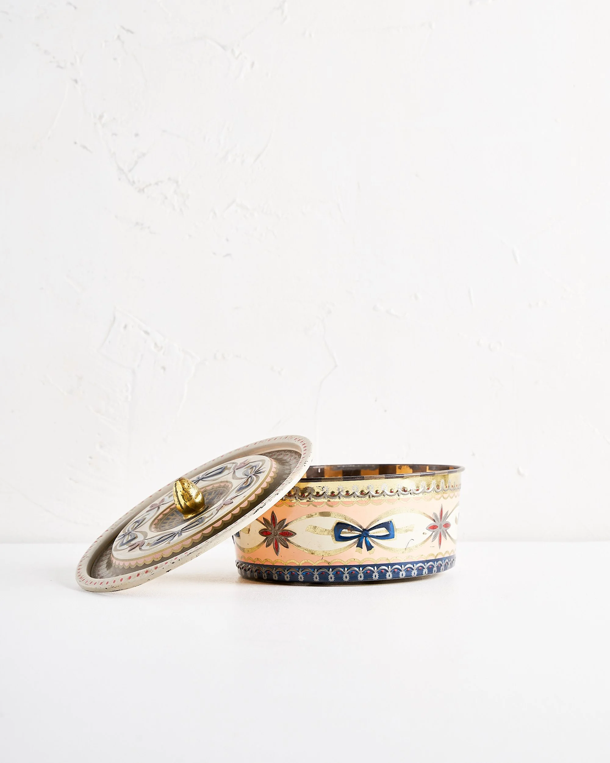 Decorative round tin container with floral and bow patterns, and a matching lid leaning against it, placed on a white surface with a plain white textured wall in the background.