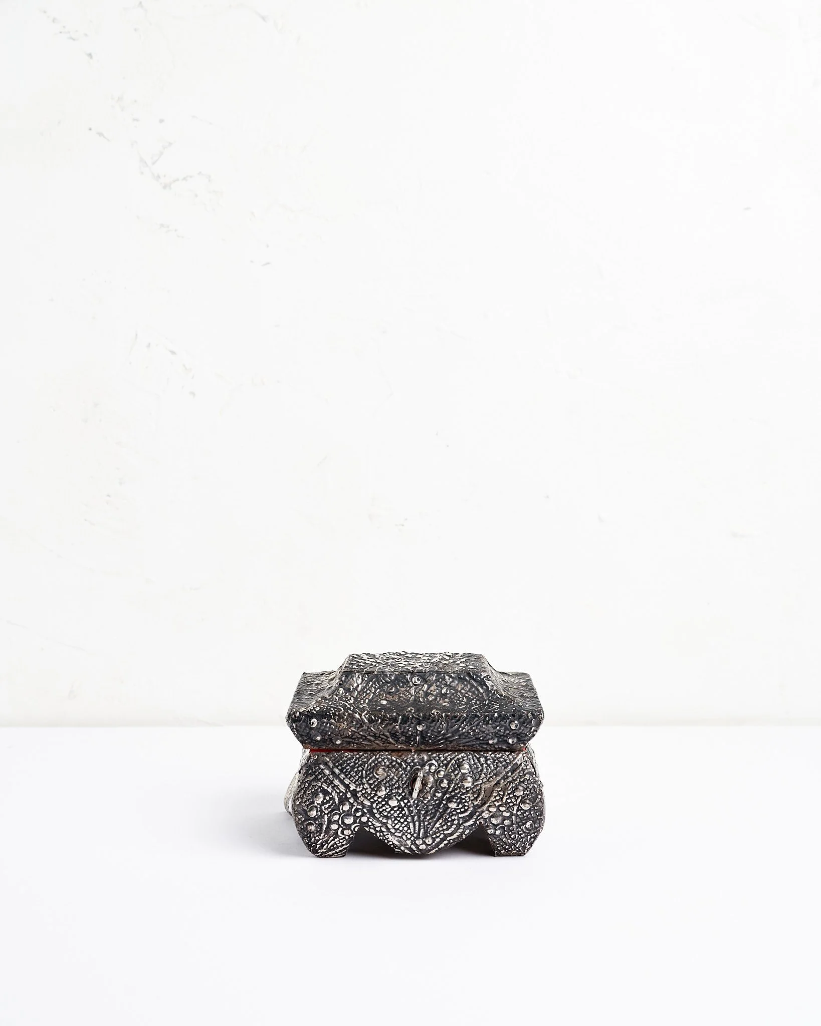 A small, intricately engraved silver metallic jewelry box with a lid resting on a plain white surface against a white background.