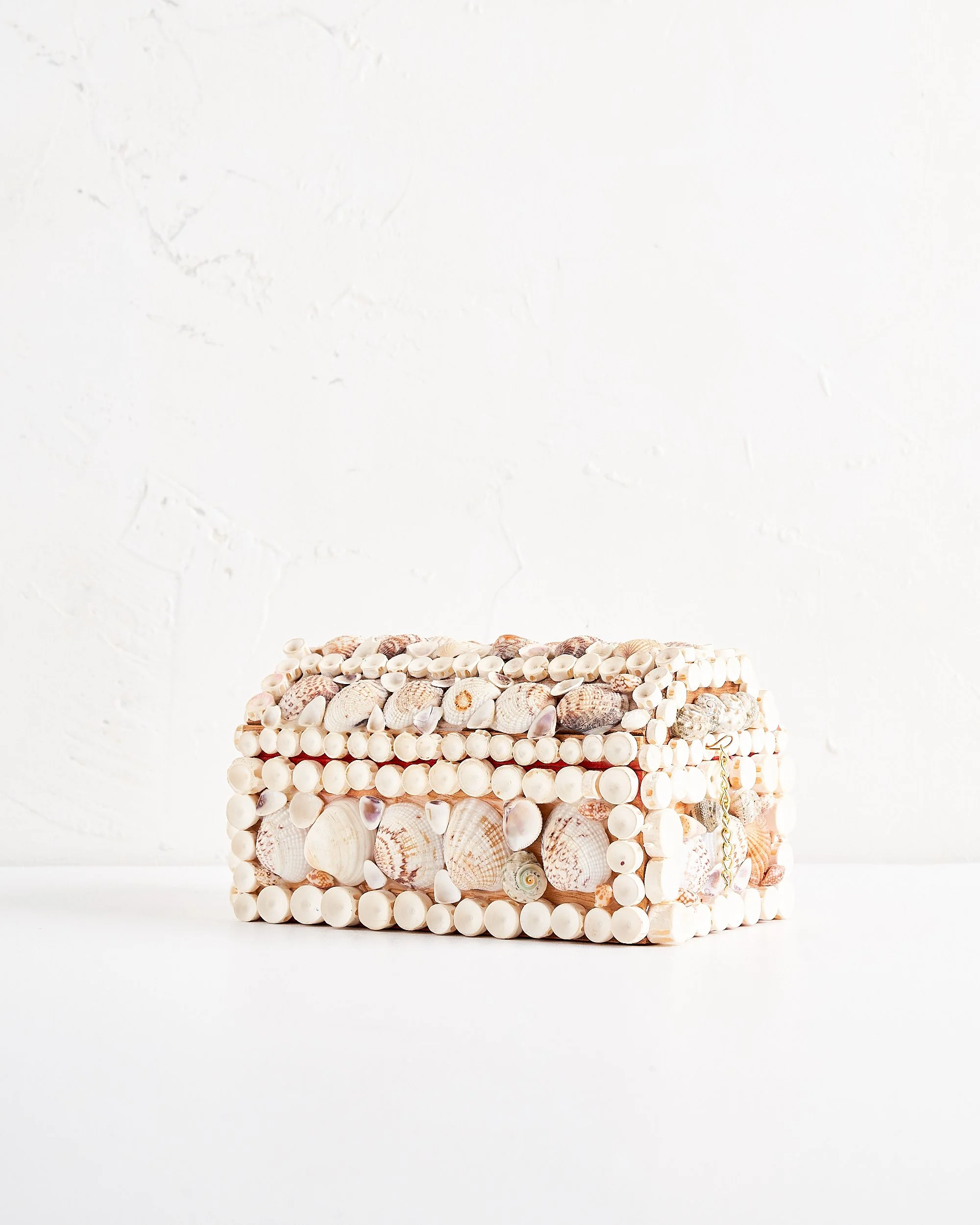 Decorative rectangular box covered in seashells with a lock on the side, placed on a white surface against a white textured background.