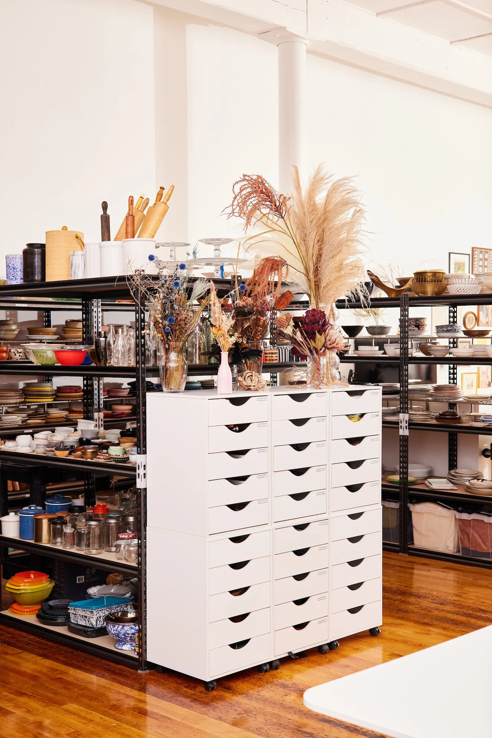 Storage room with white cabinets and black metal shelves filled with kitchenware, plates, and decorative vases.