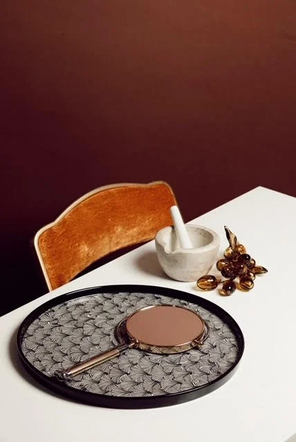 A white table with a brown and gold wooden chair, a round mirror, a magnifying glass, a white mortar and pestle, and a small cluster of dark amber stones or beads
