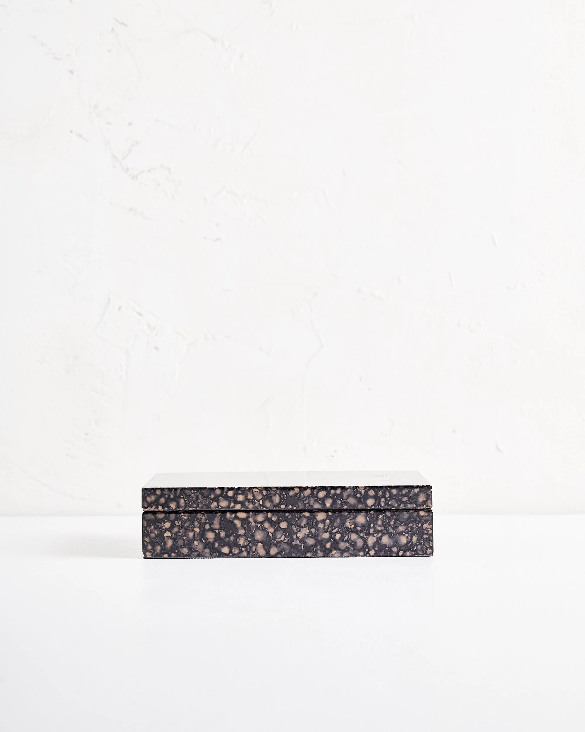 A shallow rectangular jewelry box with a black and beige speckled pattern on a white surface against a white textured wall.