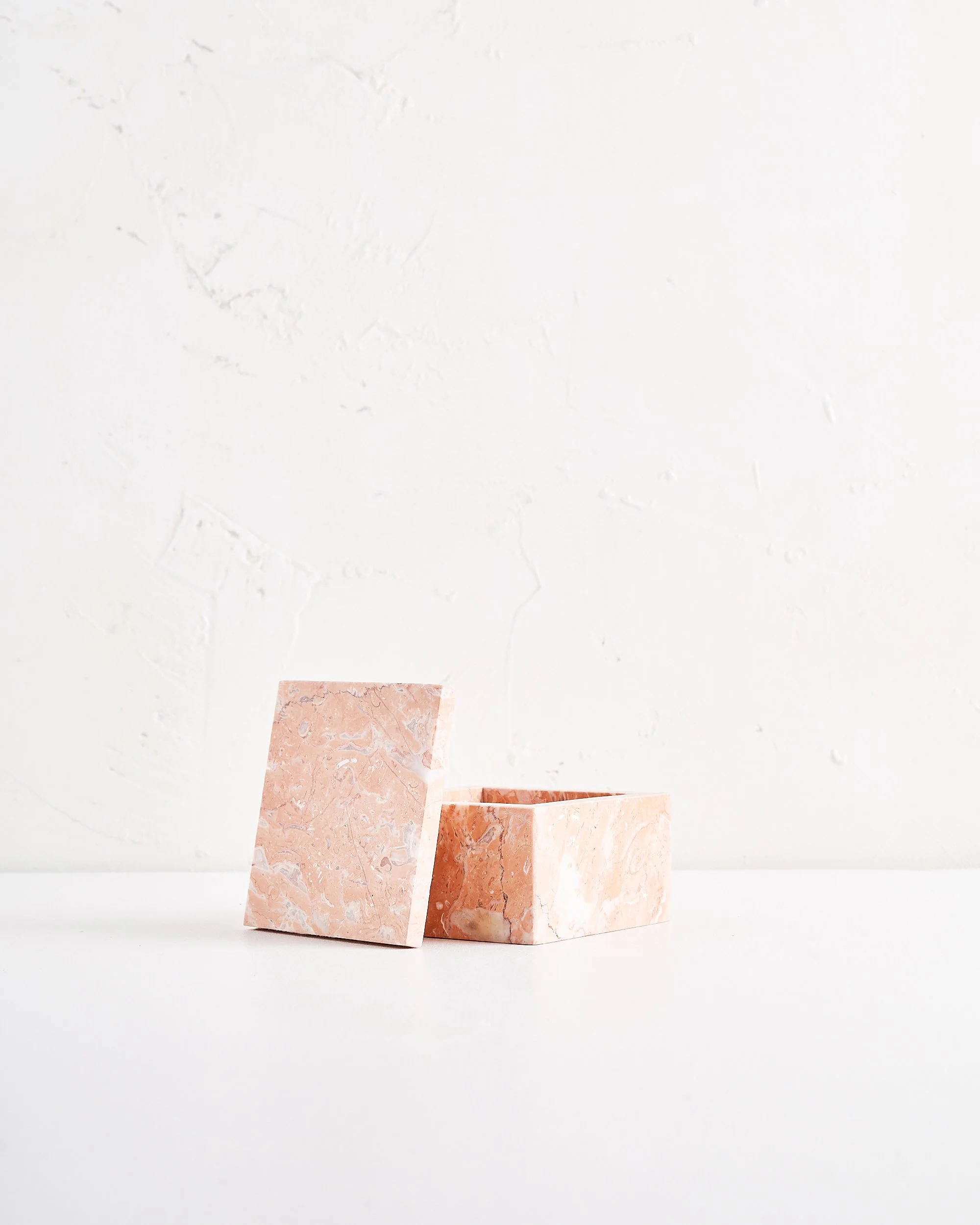 Open pink marble box, with lid leaning against it, placed on a white surface against a white textured wall.