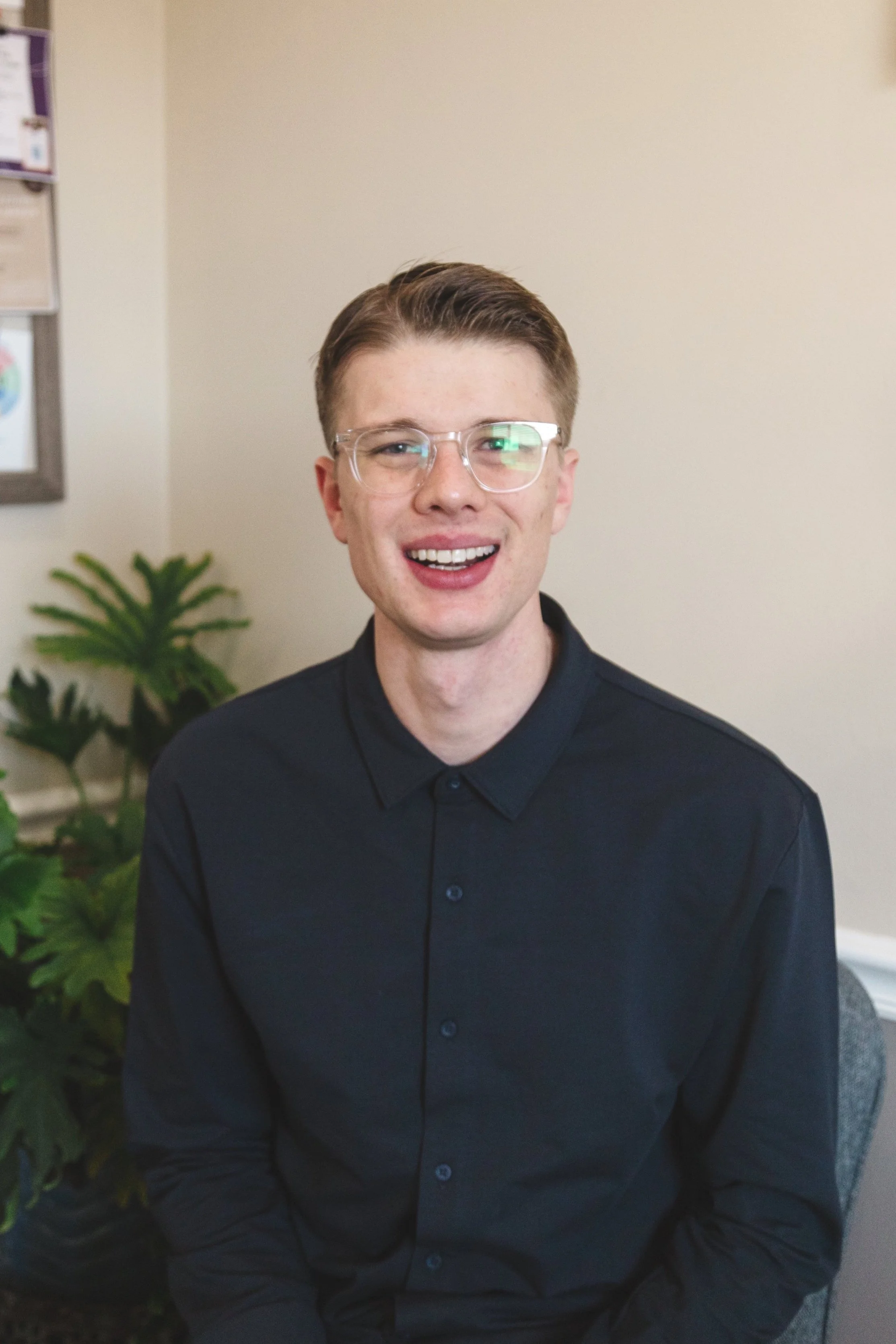 A person wearing glasses and a dark shirt smiling indoors, with plants and a wall in the background.