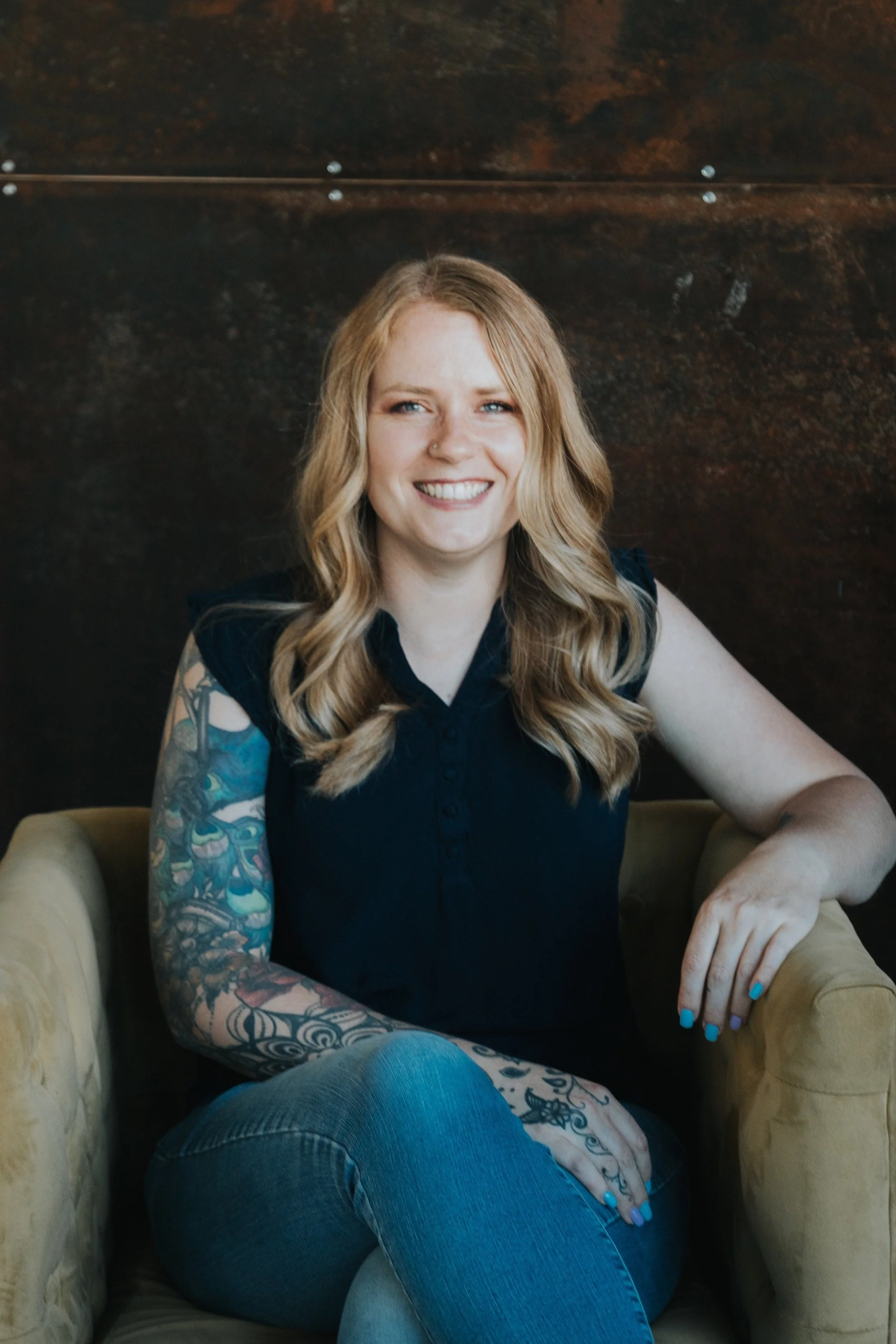 A woman with long blonde hair and glasses wearing a black top, seated indoors. She has colorful tattoos on her arm. A green plant is visible in the background.