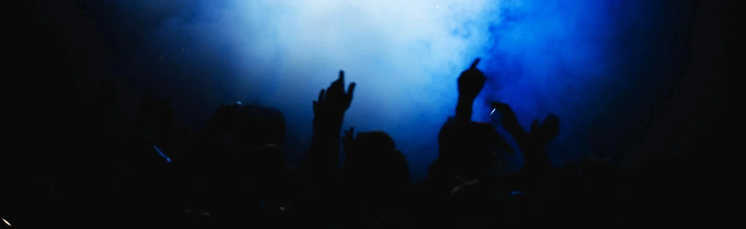 People dancing and raising their hands at a concert or nightclub, illuminated by blue stage lighting.