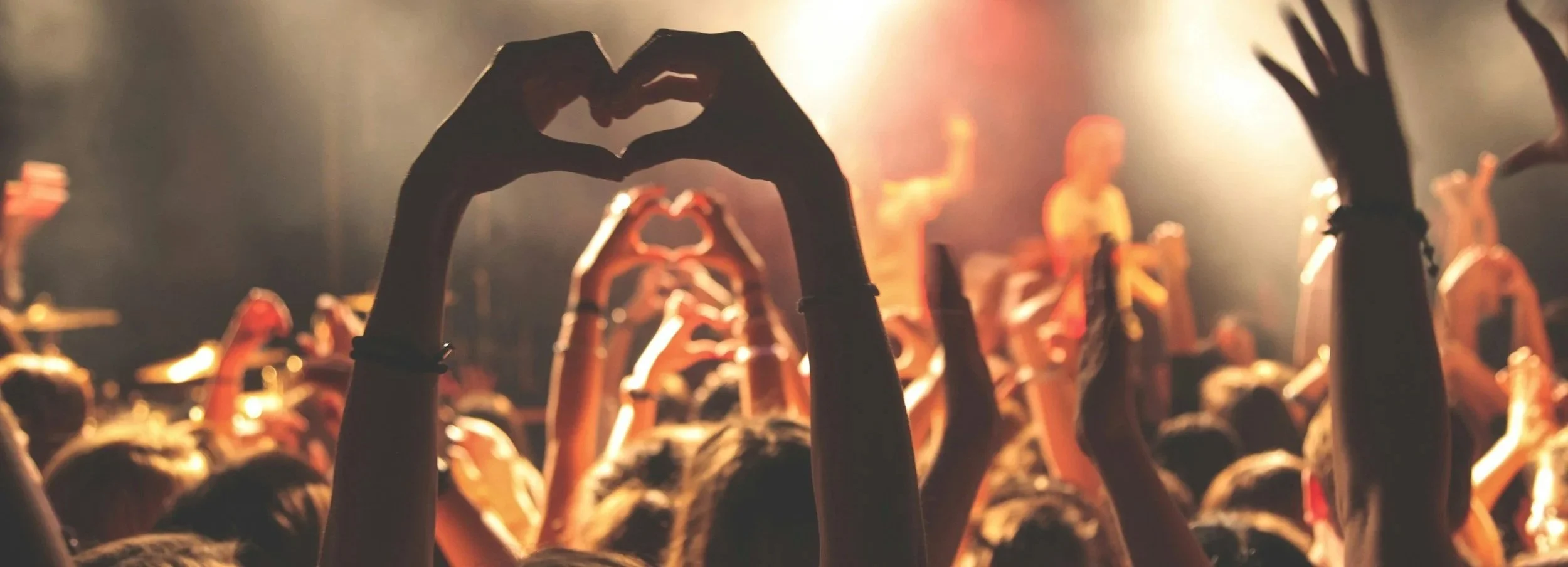 Concert crowd with people raising their hands and forming heart shapes with their fingers, stage lighting in the background.