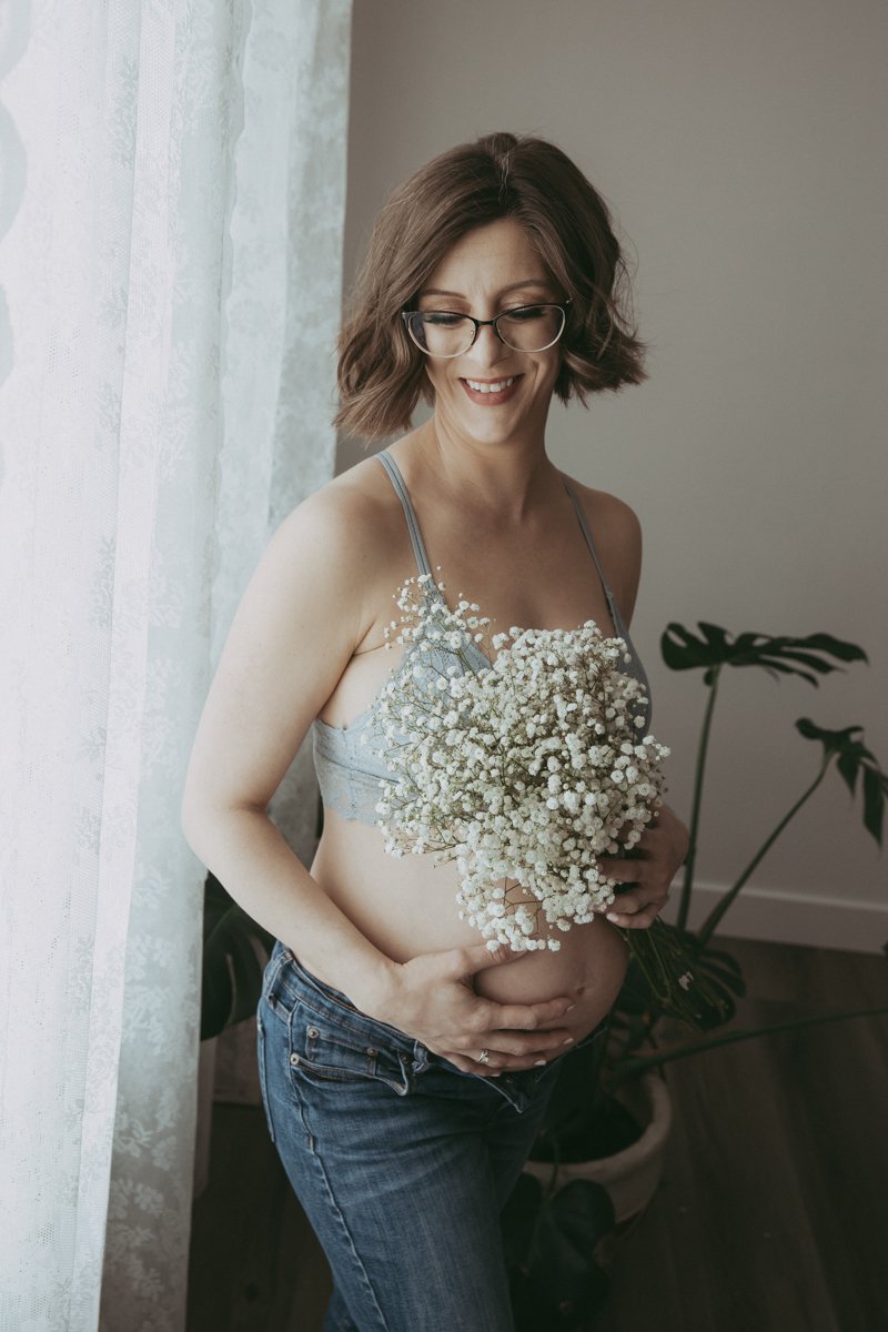 A maternity boudoir photo of a pregnant woman wearing a blue bra and jeans while holding baby's breath against herself, she is smiling and holding her belly.