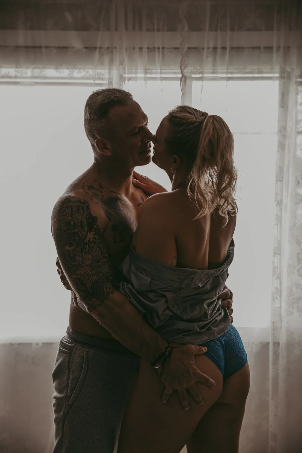 a man and woman standing in front of a window during a couples boudoir session in Edmonton