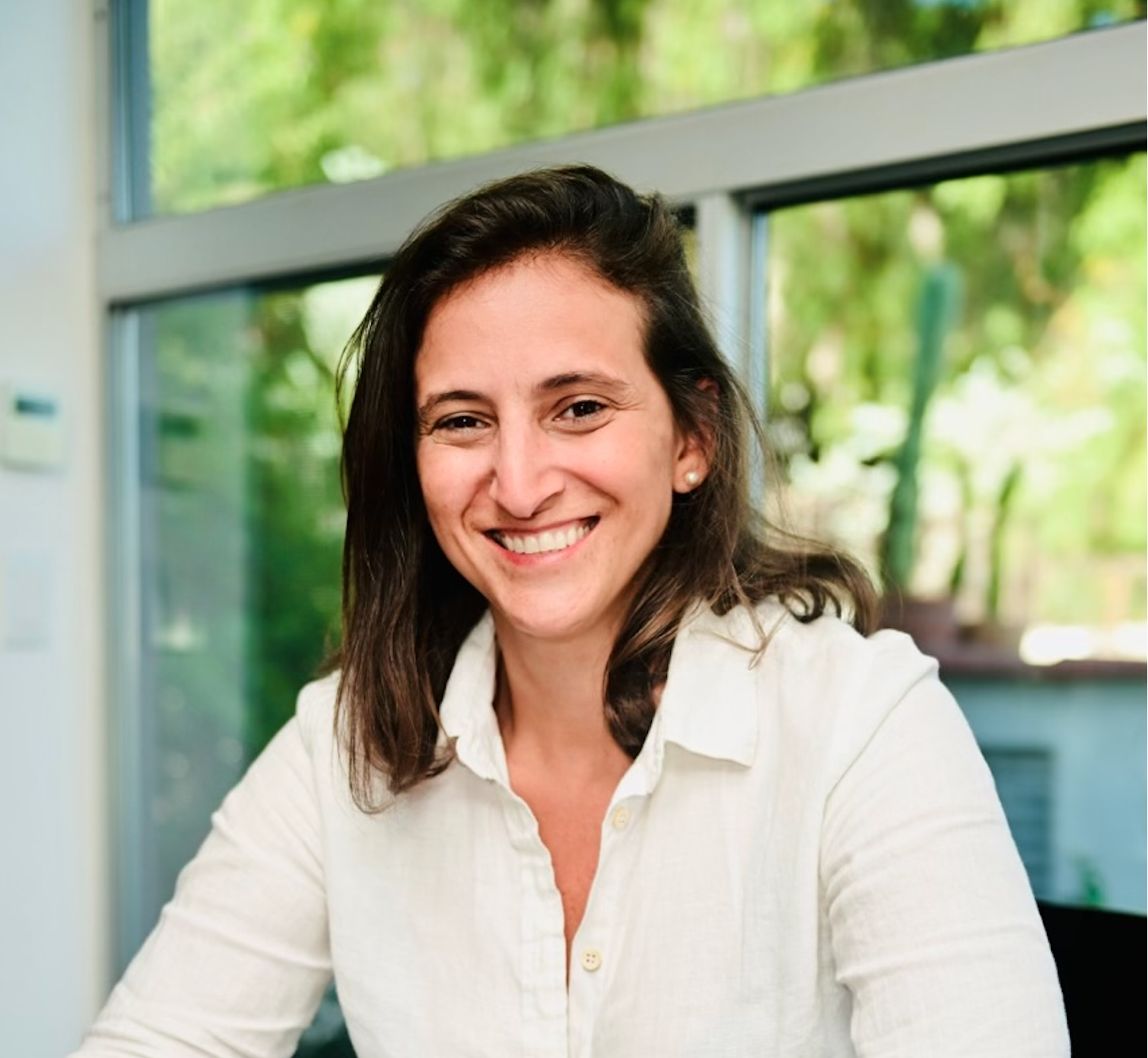 A woman with shoulder-length brown hair smiling at the camera, sitting indoors with large windows and greenery outside.