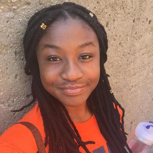 A young girl with long dreadlocks and a bright smile, wearing an orange shirt, standing against a textured wall.