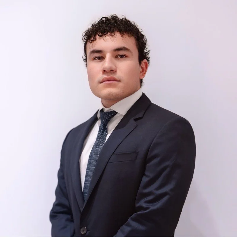 Young man in a navy suit, white shirt, and patterned tie posing against a plain white background.