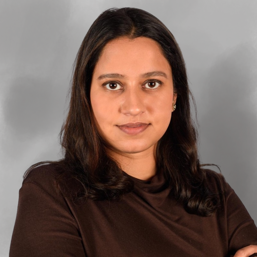 A woman with dark brown hair, wearing a dark brown top, looking directly at the camera with a neutral expression, standing against a plain gray background.