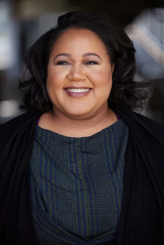 A woman with dark hair, smiling, wearing a blue patterned top and a black jacket, standing outdoors with a blurred background.