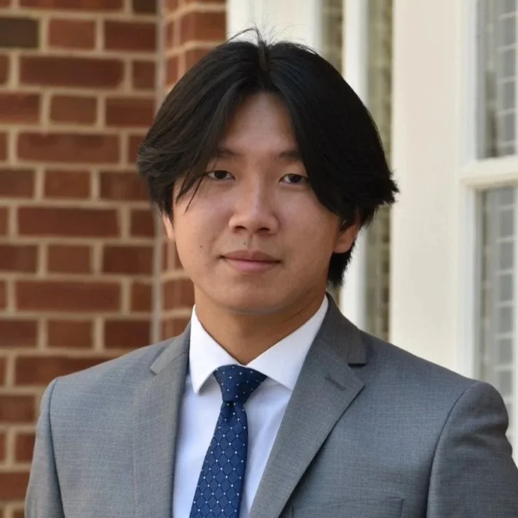 A young man with black hair, wearing a gray suit and a blue tie with white dots, standing outdoors in front of a brick wall and a window.