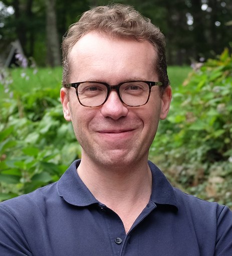 A man with glasses and curly hair smiling outdoors, standing in front of green foliage.