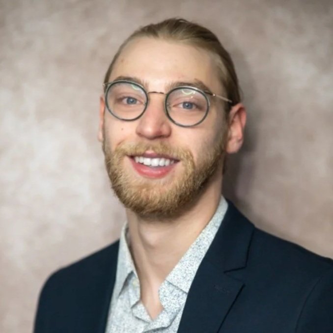A young man with sunglasses, a beard, and a mustache, smiling at the camera, wearing a suit jacket and a patterned shirt, standing against a neutral background.