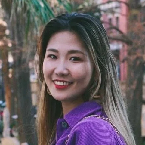 A young woman with long hair smiling outdoors, wearing a purple jacket.