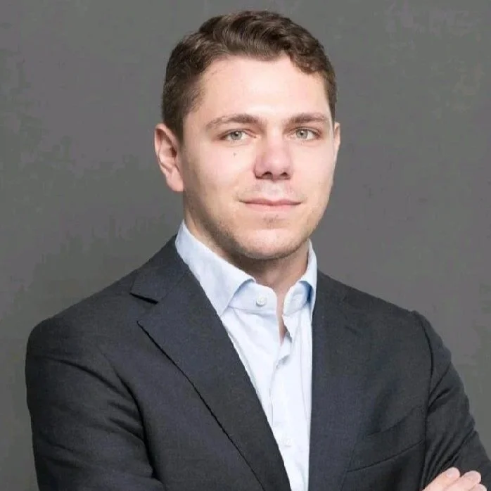 A young man with short brown hair wearing a dark suit jacket and light blue collared shirt, standing against a gray background.
