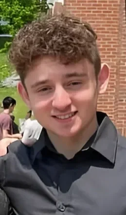 A young man with curly brown hair, wearing a dark gray shirt, smiling outside near a brick building.