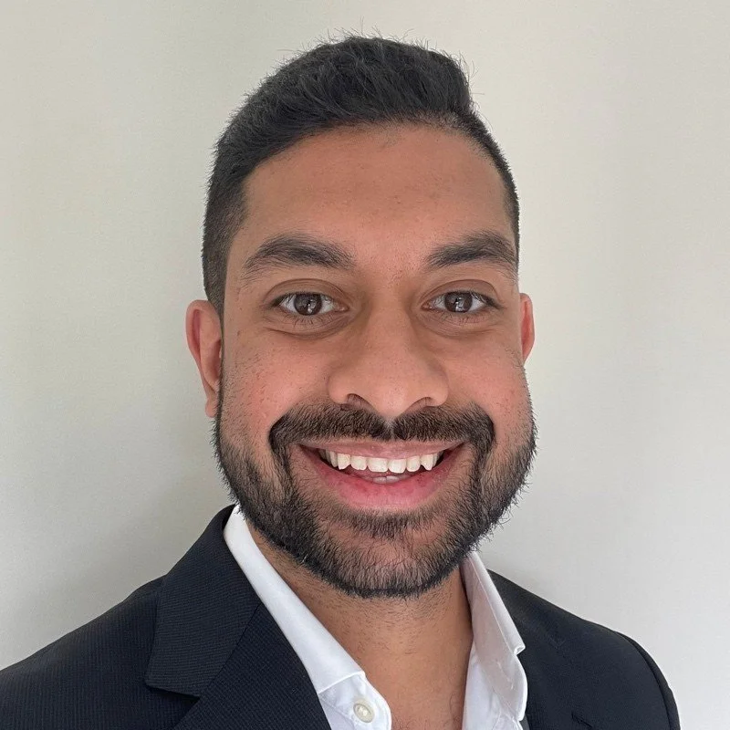 Close-up of a smiling man with dark hair, beard, and mustache, wearing a black suit and white shirt against a light-colored background.