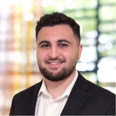 A young man with dark hair and a beard, wearing a suit jacket and white shirt, smiling in front of a colorful, blurred background.