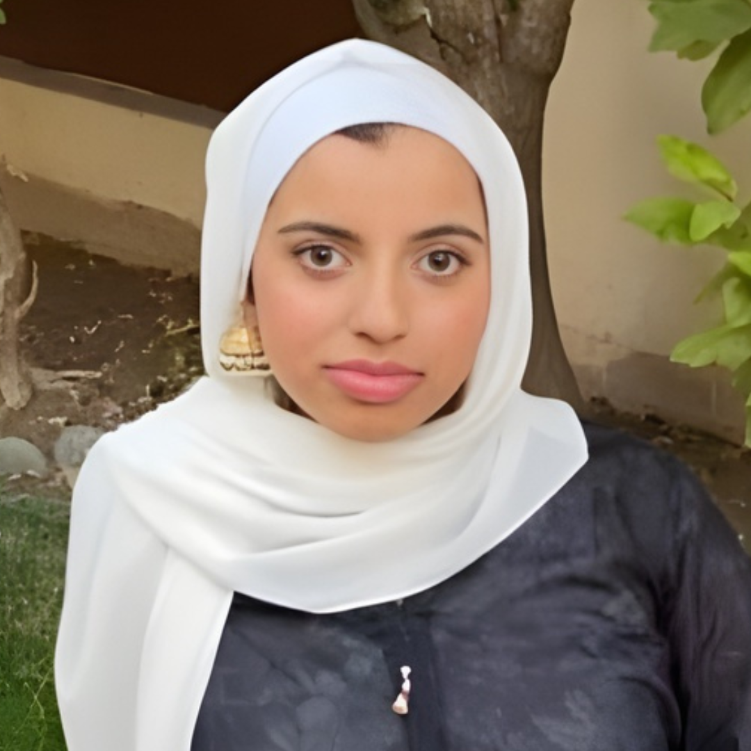 A woman wearing a white hijab and black top taking a selfie outdoors, with a tree trunk and greenery in the background.