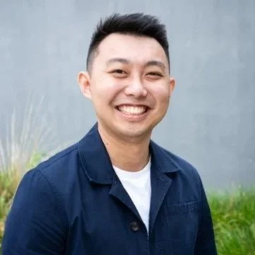 A smiling young man with short black hair, wearing a dark blue jacket over a white shirt, standing outdoors with a gray wall and some greenery in the background.