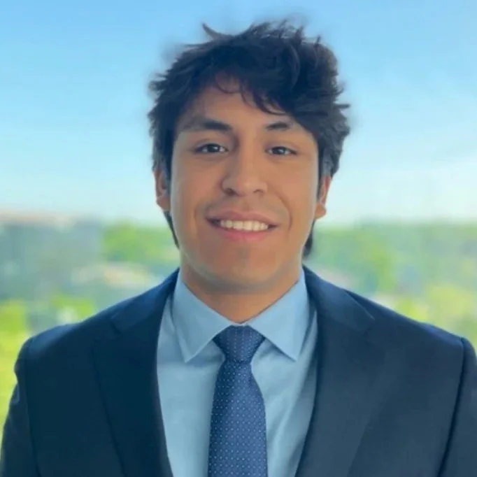 A young man in a blue suit and tie smiling in front of a window with a blurred outdoor background.
