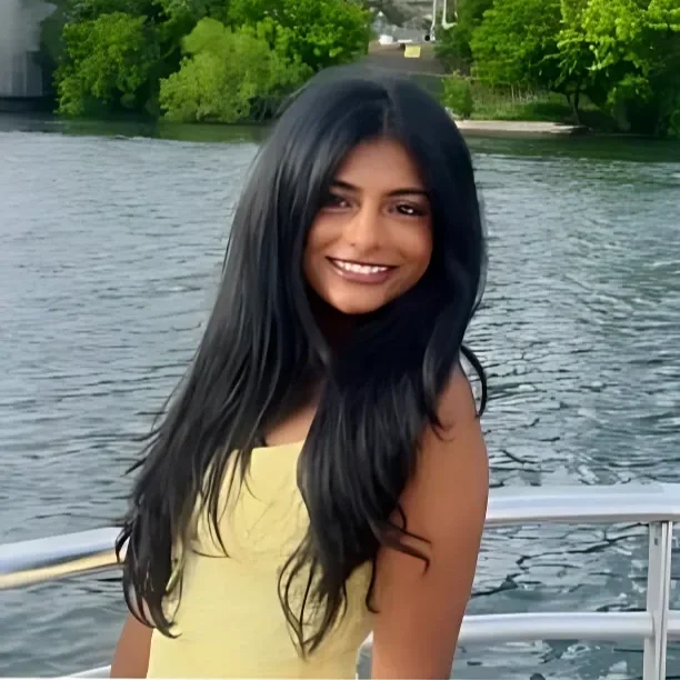 A woman with long black hair and a yellow top smiling on a boat, with water and green trees in the background.