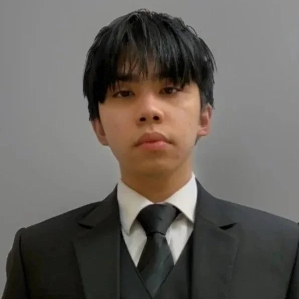 Young man in formal black suit, white shirt, and black tie standing against a plain gray background.